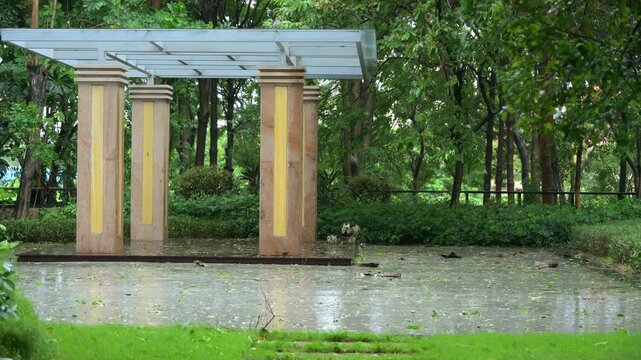 Modern stone‑column pergola standing amid wind‑driven rain inside park with wet glistening pavement covered by scattered wind‑blown fallen leaves and dense green foliage video with sound
