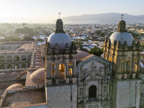 Aerial view of the historic Church of Santo Domingo de Guzman with its iconic tiled domes and twin bell towers illuminated by soft sunrise light in Oaxaca, Oaxaca, Mexico.