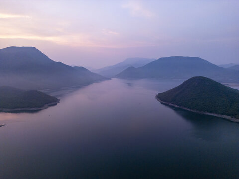Aerial view of a serene reservoir surrounded by lush green hills under a soft purple and orange sunrise sky in Jalapa, Oaxaca, Mexico.
