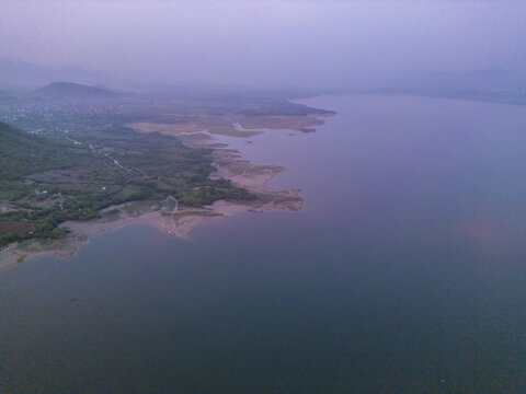 Aerial view of a serene lake at sunrise, with soft light illuminating the shoreline, lush green hills, and calm water in Jalapa, Oaxaca, Mexico.