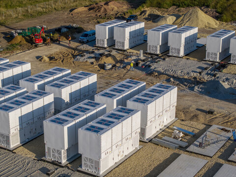 Aerial view of a battery energy storage system construction site featuring rows of white container units, construction equipment, and workers on a gravel ground in Poland.