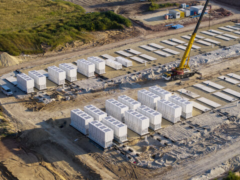 Aerial view of a battery energy storage system construction site with rows of white storage containers, a yellow crane, and construction equipment on a dirt field in Poland.