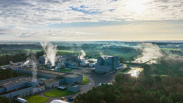 Aerial timelapse shows steam rising from an industrial plant's chimneys as morning sunlight flares over surrounding green countryside