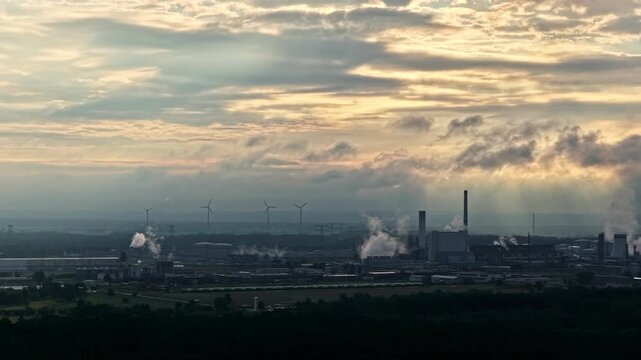 PCK Raffinerie GmbH refinery emits steam beneath layered morning clouds with distant wind turbines on the horizon.