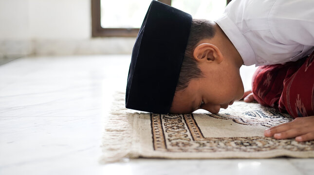 Young Muslim boy in prayer cap praying on a rug, close-up on face bowing down during Salah.