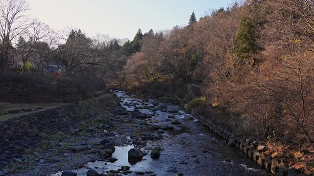 Low river flow reveals exposed rocks and a shallow riverbed surrounded by leafless trees and dry vegetation during the colder season in rural Japan.