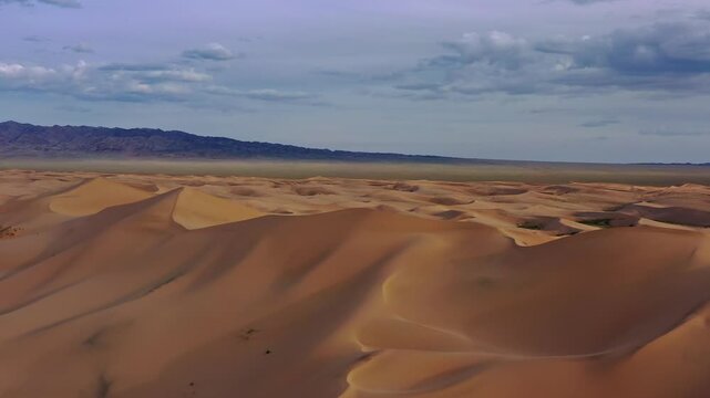 Aerial view of the vast sand dunes of the Gobi Desert with distant mountains under a cloudy sky in Mongolia.
