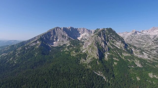 Aerial view of Bobotov Kuk peak in Durmitor National Park with rocky slopes and pine forests under a clear blue sky in Zabljak Municipality, Montenegro.