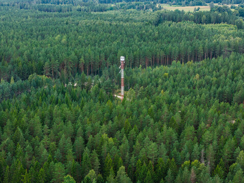 Aerial view of a forest fire observation tower with solar panels, wind turbine, cameras, and antennas rising above a dense pine forest in Talsi Municipality, Latvia.
