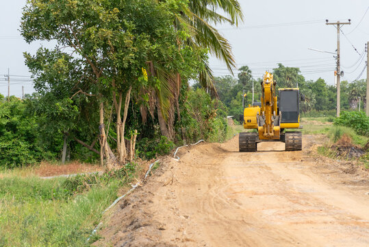Yellow excavator stands rural dirt road under construction surrounded by tropical trees grass utility poles farmland showing infrastructure development