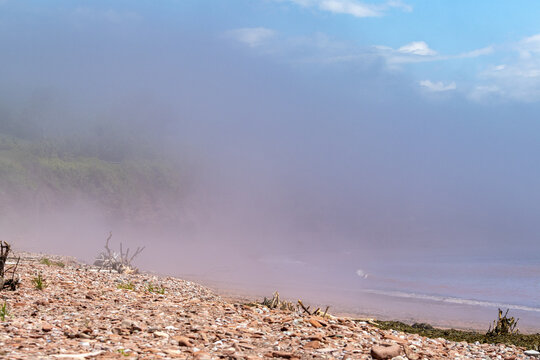 Gaspesie, Canada, 17 June 2026 : Thick reddish-purple fog rolls over a rocky beach with driftwood under a partly cloudy sky