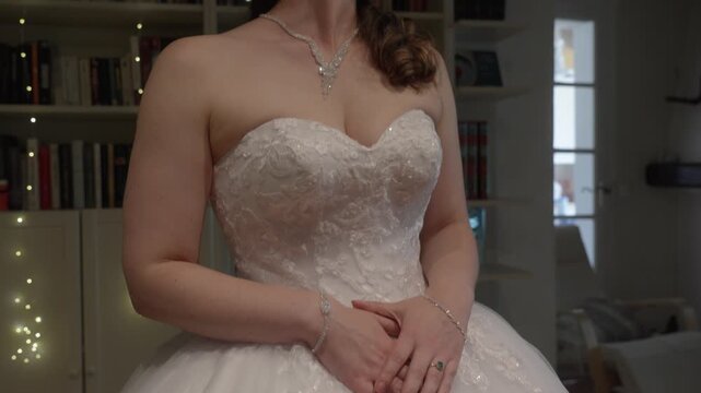 Bride in white wedding dress stands indoors with hands clasped, elegant jewelry visible