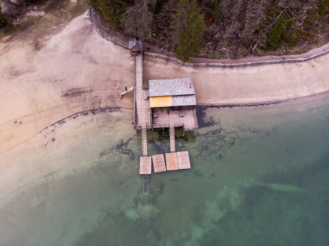 Aerial view of the wooden boathouse and pier extending into the clear turquoise waters of Lake Braies, surrounded by a sandy shoreline and pine forest in Braies, Trentino-Alto Adige, Italy.