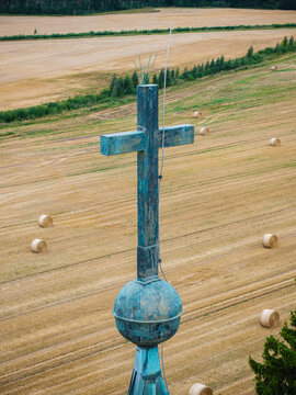 Aerial view of the copper cross and orb atop the Arlava Evangelical Lutheran Church overlooking a rural field with hay bales in Valdemarpils, Talsi Municipality, Latvia.