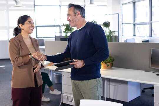 Diverse coworkers shaking hands while holding tablet at open office desk with monitors and plants