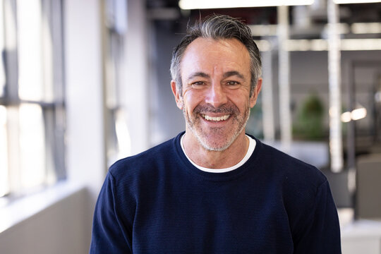 Senior male smiling while standing in modern open-plan office with large windows and potted plants