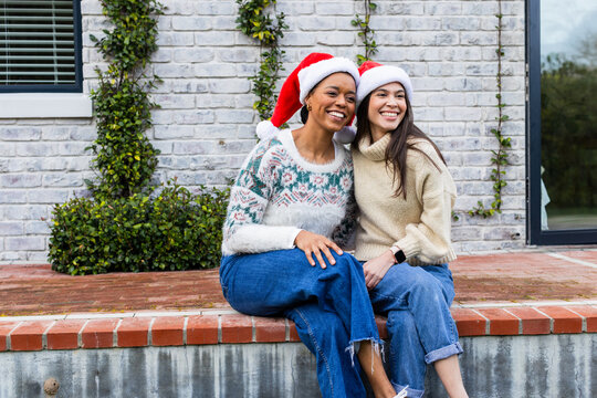 Diverse female friends sitting on brick ledge wearing Santa hats and sweaters leaning together
