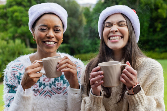 Diverse mother and daughter standing in garden holding ceramic mugs wearing Santa hats and sweaters
