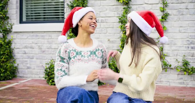 Two women 20s,30s on patio ledge laughing, leaning together and adjusting Santa hats for Christmas