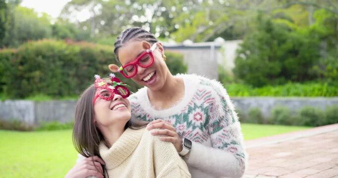 Two women 20s and 30s laughing and leaning on brick ledge in yard adjusting Christmas glasses