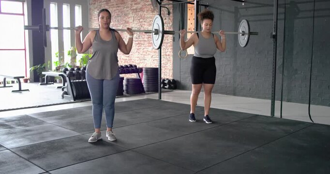 On cue, Diverse female friends doing lunges and pressing barbells overhead in gym, then high-fiving