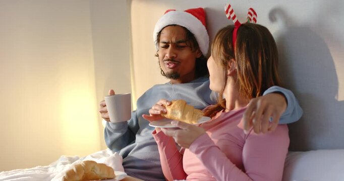 Diverse couple wearing festive headwear tasting croissant on bed tray sipping mug and wiping crumbs
