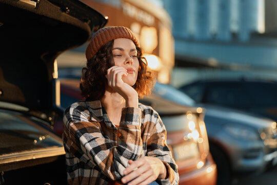 Woman car urban portrait candid golden hour glow: a thoughtful woman leaning on a car trunk in a city street, plaid jacket and beanie, authenticity and mindful living, emotional storytelling mood.