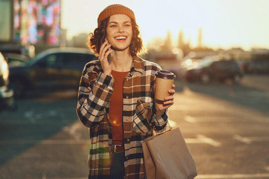 Woman holding coffee and shopping bag, smiling while on phone in sunlight in an urban parking lot at sunset. Candid lifestyle scene with authenticity, golden hour glow, mindful living and emotional