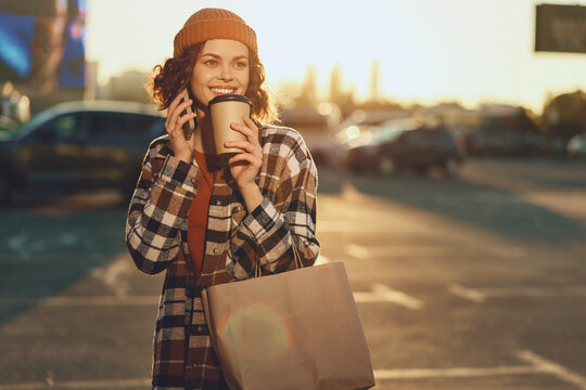 Woman with coffee and phone holding a shopping bag outdoors in urban street at golden hour, smiling candidly with authenticity and a golden hour glow, mindful living.