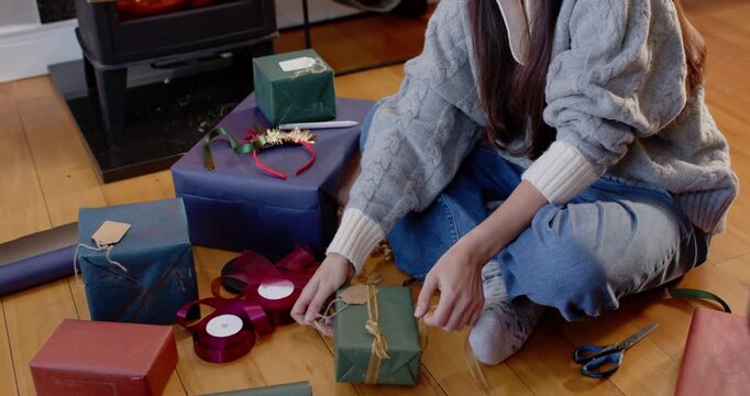 Woman in chunky knit sweater tying twine on green gift for Christmas beside wood-burning stove