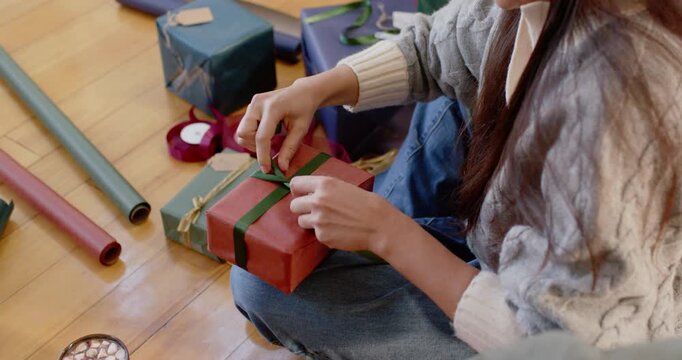 Woman sitting on floor reaching for green-ribbon, tying bow on red gift for Christmas, copy space