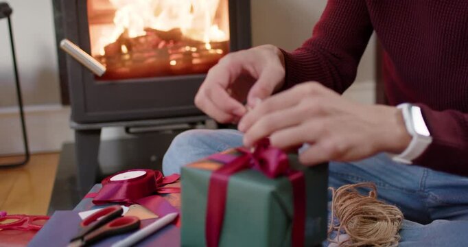 Man responding to wood stove glow, tying twine around red ribbon on Christmas gift, copy space