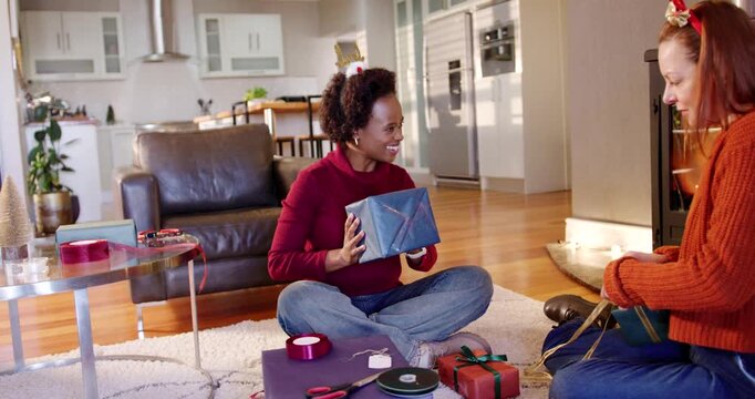 Diverse women on living room rug wrapping Christmas gifts, holding blue box and tying gold bow