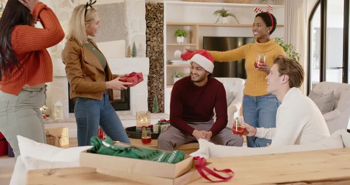 Diverse friends presenting red gift box, placing Santa hat and toasting Christmas in living room