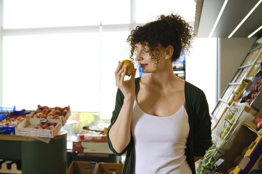 Woman stands in market holding lemon to her face. She looks thoughtful while surrounded by various fruits and vegetables. Natural light comes through windows illuminating the space