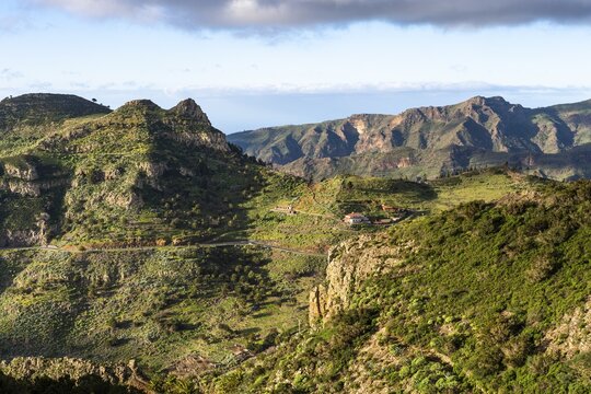 Landscape in the south-east of La Gomera. Some houses in the village of Vegaipala. Mountain slopes with palm trees and agaves, a road, sun and clouds. La Gomera, Canary Islands, Spain