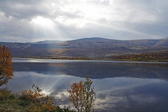 A lake with mountains in the background, rays of light penetrate the clouds, autumn leaves and their colours are visible, Valasjoen, Hjerkinn, Dovrefjell-Sunndalsfjella National Park, Norway