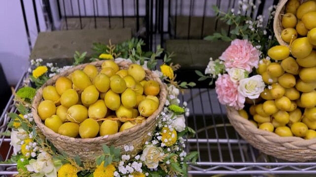 Close-up of fresh yellow dates beautifully arranged in a woven wicker basket, decorated with pink and white flowers. An elegant indoor cultural food display resting on a metal shelf.
