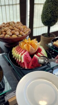 Close-up vertical shot of a person using metal tongs to serve a slice of fresh watermelon onto an empty white plate. The healthy fruit platter sits at an indoor restaurant catering buffet station.