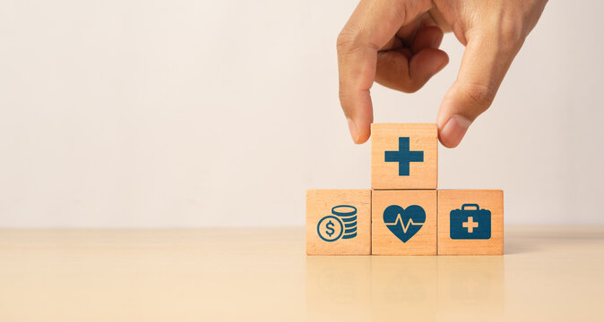 Hand placing a wooden block with a medical cross symbol on top of other blocks featuring healthcare, finance, and insurance icons, representing health services, medical support, and financial coverage