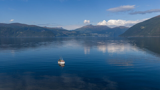Aerial view of a small fishing boat floating on the calm blue waters of a fjord, surrounded by majestic mountains in Liabygda, More og Romsdal, Norway.