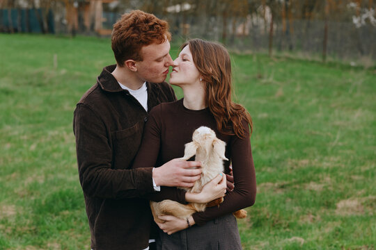 A couple embraces outdoors with a small goat kid between them in a grassy field, conveying warmth, affection, and rural lifestyle. A gentle moment captures love, care, and connection in a casual farm