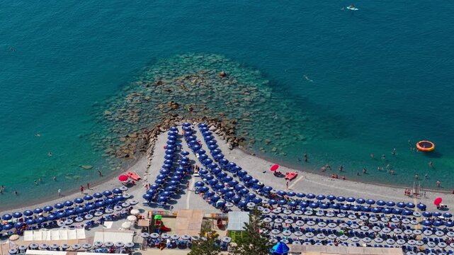 Drone view over a busy summer beach in Bordighera, with rows of blue umbrellas, people swimming in turquoise Mediterranean water and rocks visible beneath the clear sea.
