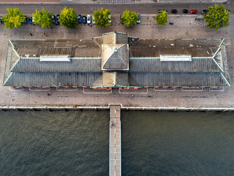 Aerial view of the historic Fischauktionshalle building along the waterfront with a wooden pier extending into the Elbe river, surrounded by trees and cobblestone paths in Hamburg, Hamburg, Germany.