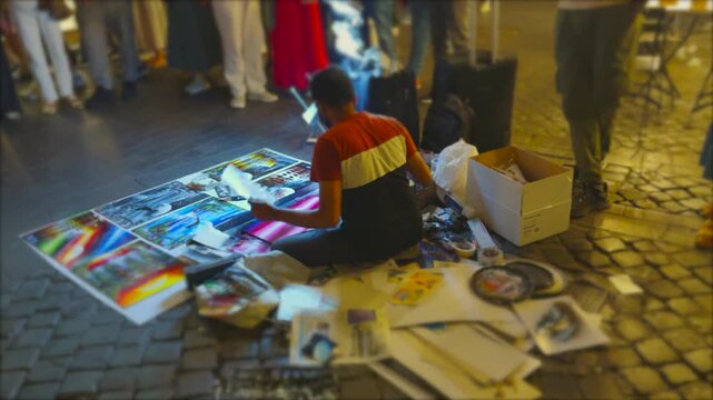 Street Artist Creating Spray Paint Art at Night Surrounded by Crowd and Displayed Prints in Rome Italy