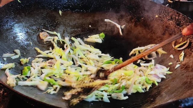 Chopped cabbage onion and green capsicum being vigorously stir fried in a hot wok with steam rising during street food preparation