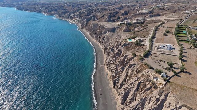 Tall eroded bluffs edge a narrow dark beach and clear sea beside scattered farm fields and villas as the camera tracks along the coast.