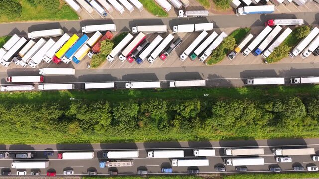 Aerial perspective of a truck parking area filled with trucks and cars, illustrating the ongoing movement of vehicles along the road and surrounding greenery