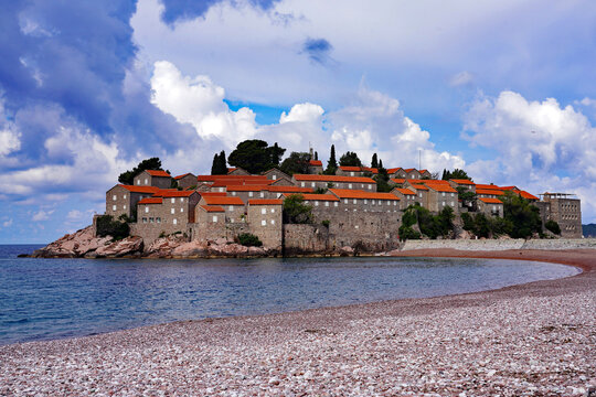 The fortified island village of Sveti Stefan overlooks a curved pink pebble beach and the turquoise waters of the Adriatic Sea under a dramatic cloudy sky in Montenegro