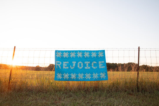 hand made quilt that says rejoice hanging from rural fence at sunset 